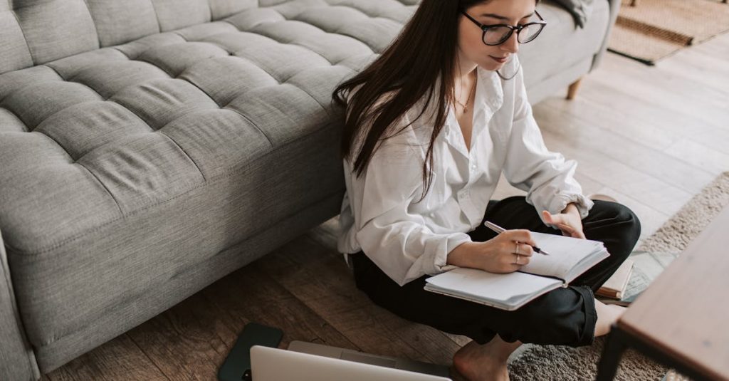 A young woman sitting on the floor using a laptop and writing notes in a cozy apartment.
