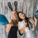Two women in festive outfits holding balloons at a party with foil curtains.